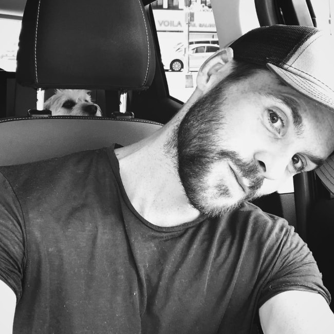 Tom Riley in a black-and-white car-selfie with his dog in the background in September 2018