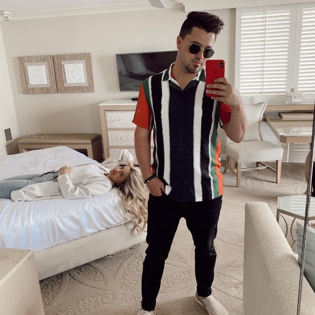 Kyler Fisher as seen while taking a mirror selfie with wife Madison Nicole Fisher lying on the bed in the background in Laguna Beach, Orange County, California, United States in April 2019