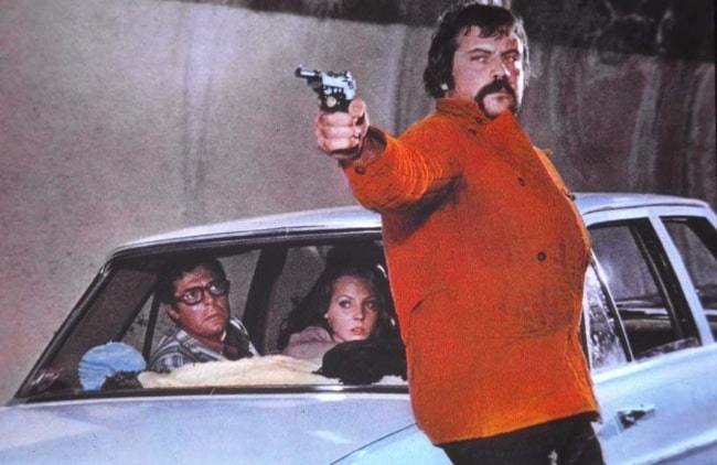 Oliver Reed (in red) as seen along with Marcello Mastroianni and Carole André (sitting inside the car) in a scene shot on the set of the film Mordi e fuggi (1973) by Dino Risi
