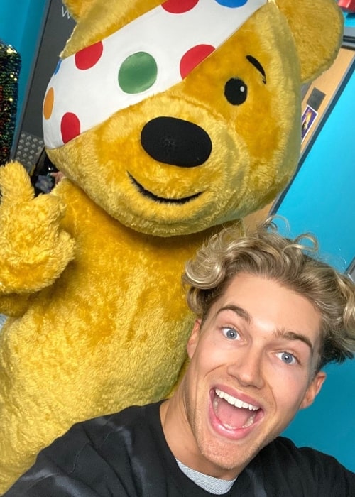 AJ Pritchard as seen in a selfie with a teddy bear mascot costume posing at the back of him in November 2019