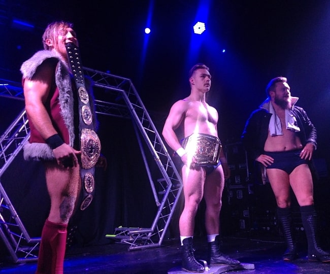 Tyler Bate (Center) as seen while holding the WWE United Kingdom Championship belt and standing on the Progress Tag Team Championship shield in January 2017