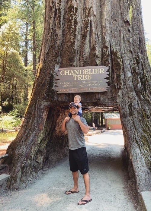Tanner Tolbert at the Drive-Thru Tree Park having fun standing under a big tree with a small baby in June 2020