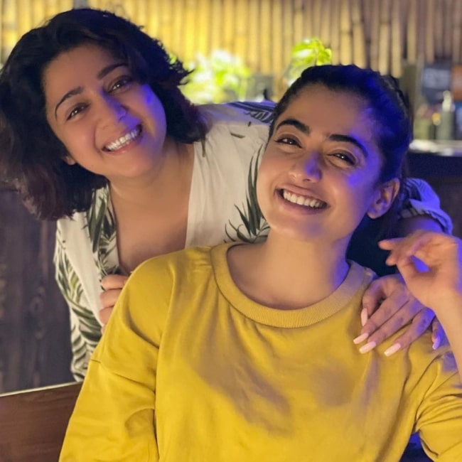 Charmy Kaur posing from behind Rashmika Mandanna as they both smile for the camera